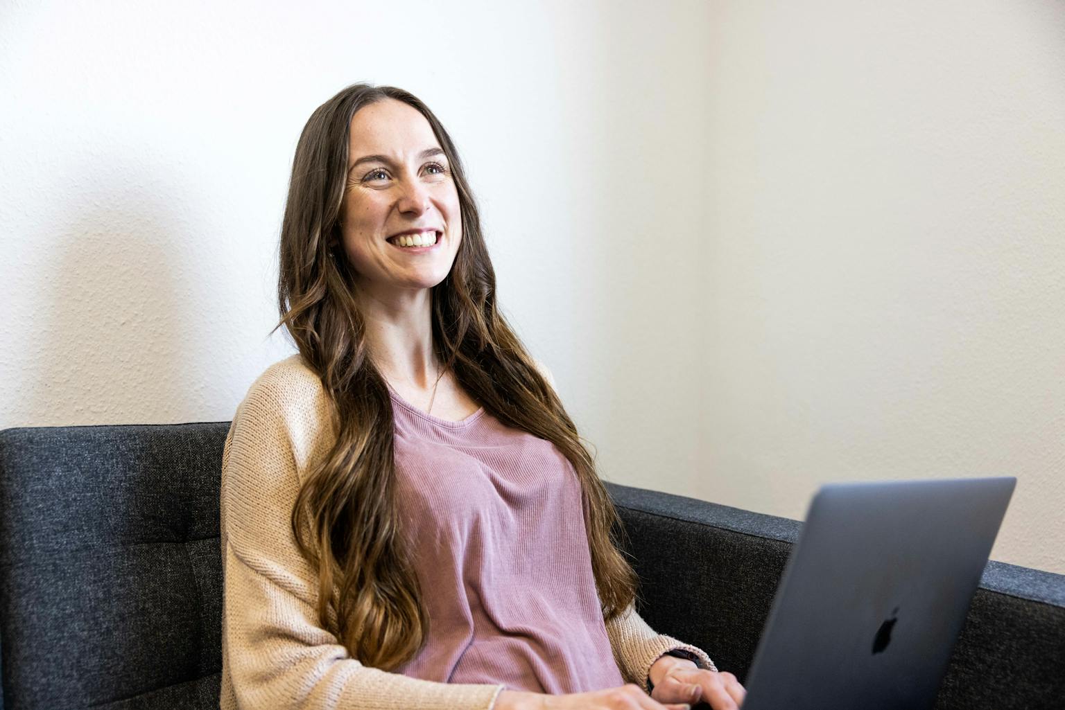 Eine junge Frau mit langen Haaren sitzt mit ihrem Laptop auf dem Sofa und grinst freundlich.